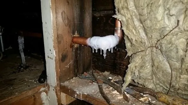 A copper elbow coated in frost with icicles hanging from it beside torn wall insulation