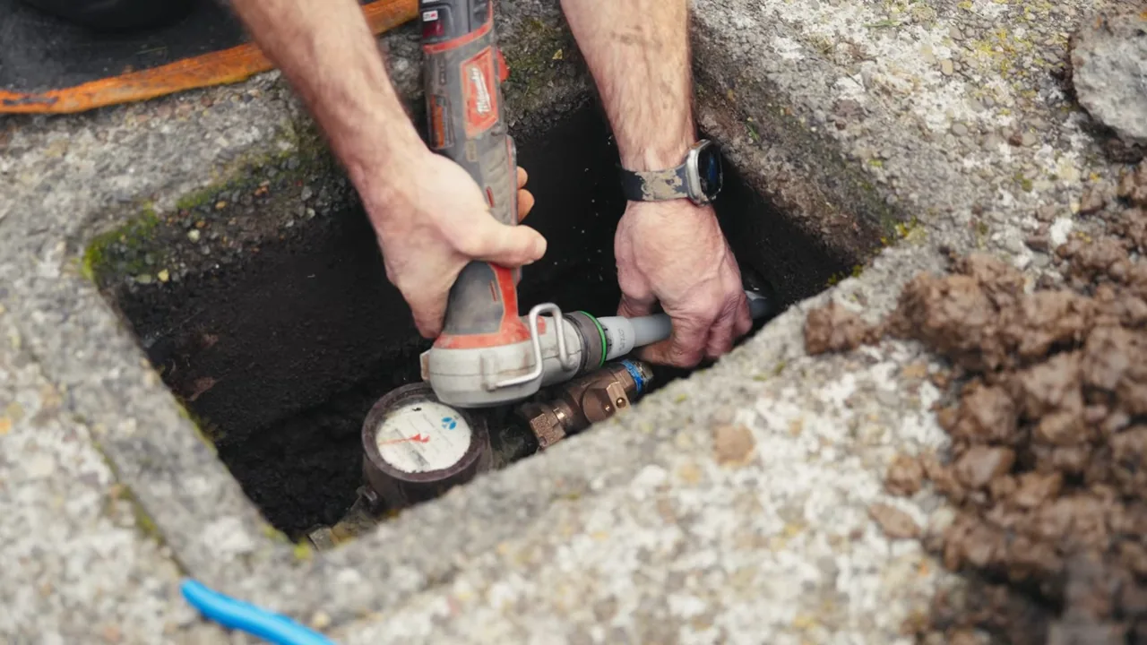 Hands holding a power tool on a pipe fitting next to a water meter set in a block ground box